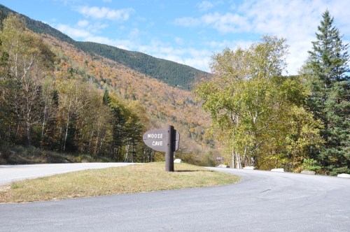 A sign for Moose Cave near a winding road, surrounded by trees and mountains with autumn foliage.