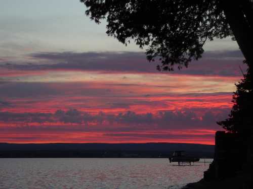 A vibrant sunset over a calm lake, with pink and purple clouds and a silhouette of a boat docked nearby.