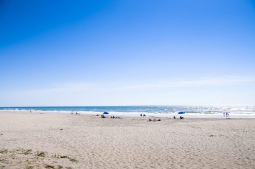 A sunny beach scene with a clear blue sky, gentle waves, and a few people relaxing under umbrellas.