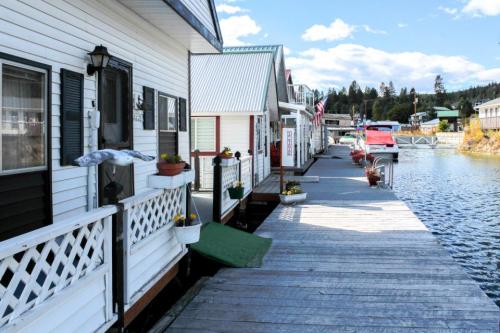 A scenic view of houseboats along a wooden dock, with potted plants and a calm waterway under a blue sky.