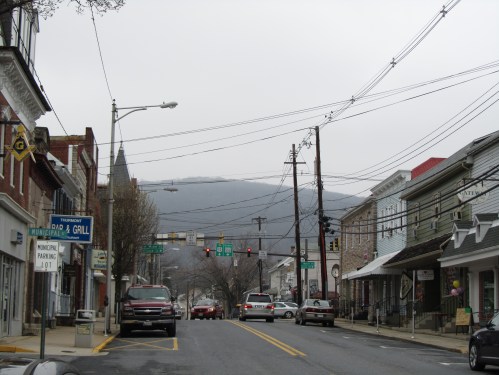 A quiet street scene with shops, power lines, and a view of mountains in the background under a cloudy sky.