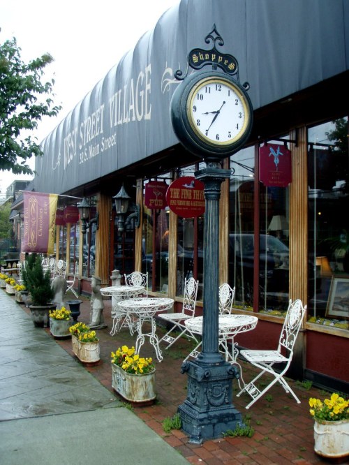 A vintage clock stands in front of a quaint shop with outdoor seating and flower planters along the sidewalk.