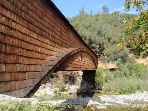 A wooden covered bridge arches over a river, surrounded by lush greenery and rocky riverbanks.