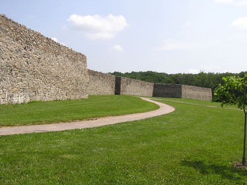 A grassy area with a winding path alongside a tall stone wall under a blue sky with a few clouds.