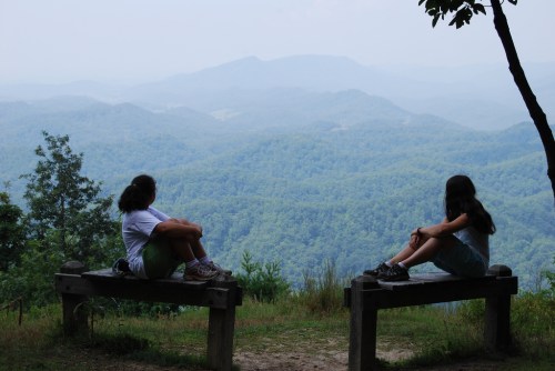Two girls sit on benches, gazing at a misty mountain landscape filled with lush green hills.