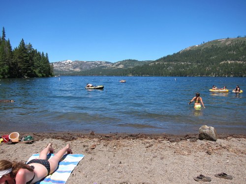 A sunny lakeside scene with people swimming and relaxing on the beach, surrounded by trees and mountains.