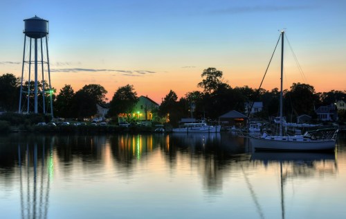 A serene waterfront scene at dusk, featuring a water tower, boats, and colorful reflections on calm water.