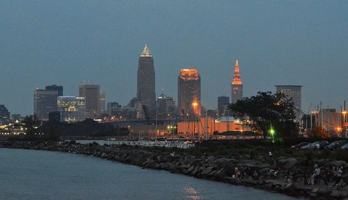 Cleveland skyline at dusk, featuring illuminated skyscrapers and a calm waterfront with boats and people along the shore.