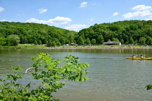 A serene lake surrounded by lush green hills, with people enjoying the beach and two kayakers paddling on the water.