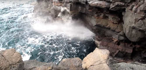 Waves crashing against rocky cliffs, creating mist and splashes in a coastal landscape.