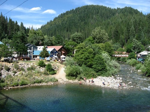 A scenic view of a riverbank with people enjoying the water, surrounded by lush greenery and quaint houses.