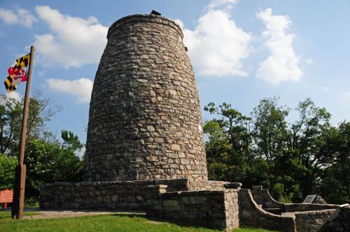 A tall stone tower surrounded by greenery, with a Maryland flag nearby and a clear blue sky above.