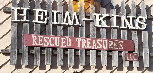 Sign on a wooden fence reading "Heim-Kins Rescued Treasures LLC" in white and red letters.