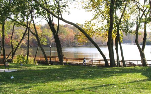 A serene lakeside scene with trees, a grassy area, and a wooden walkway along the water, showcasing autumn colors.