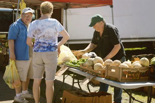 A farmer's market scene with two customers interacting with a vendor while selecting fresh produce.