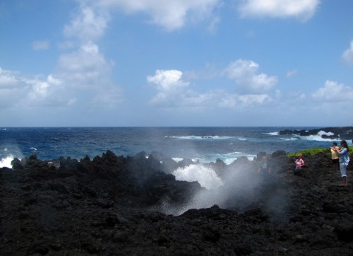 A rocky coastline with waves crashing, creating mist, under a blue sky with scattered clouds. People are observing the scene.