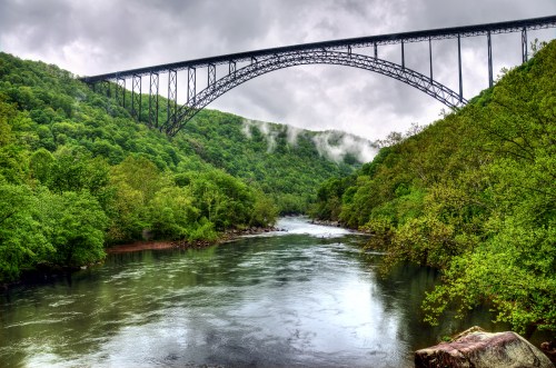 A scenic view of a river surrounded by lush green trees, with a large arch bridge spanning above it under a cloudy sky.
