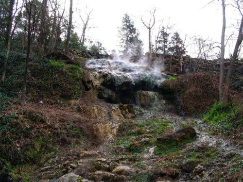 A steaming waterfall cascades down a rocky slope, surrounded by trees and lush greenery.