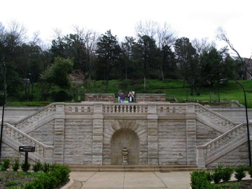 A stone staircase leads to a grassy area with trees, where three people are walking. Overcast sky above.