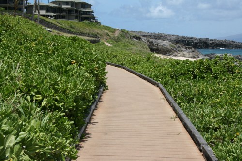 A wooden pathway winds through lush greenery, leading to a rocky beach and ocean under a partly cloudy sky.