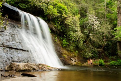 A serene waterfall cascades into a calm pool, surrounded by lush greenery and two people sitting nearby.