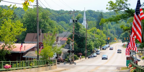 A small town street lined with trees, flags, and a church in the background, with cars driving along the road.
