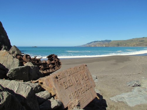 Rusty sign on rocky beach with ocean waves and hills in the background under a clear blue sky.