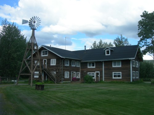 A large wooden house with multiple windows, a grassy yard, and a windmill nearby under a partly cloudy sky.