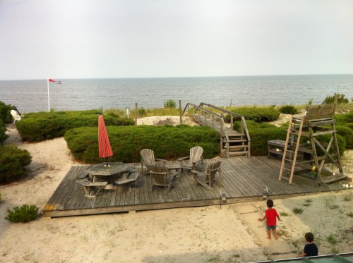 A sandy beach area with wooden chairs, a red umbrella, and a view of the ocean under a cloudy sky.