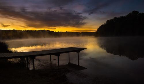 A serene lake at sunrise, with mist rising and a wooden dock extending into the calm water, surrounded by trees.