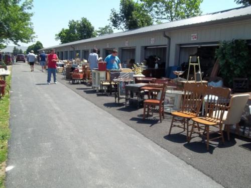 A row of storage units with people browsing various pieces of furniture displayed outside on a sunny day.