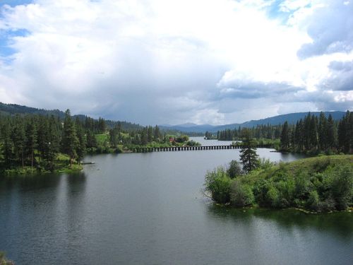 A serene lake surrounded by trees and mountains under a cloudy sky, with a wooden bridge in the distance.