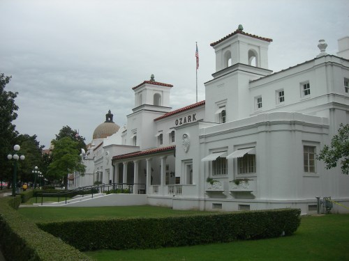 Historic white building with red-tiled roof, featuring decorative towers and a dome in the background, set in a green landscape.