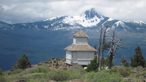 A small white building on a rocky hillside, with snow-capped mountains and cloudy skies in the background.
