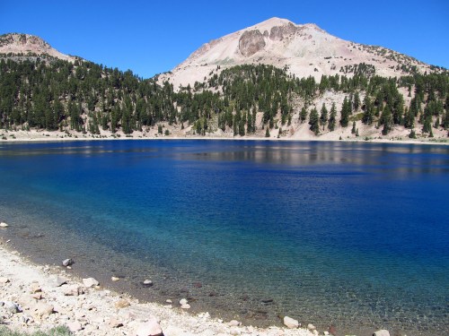 A serene lake surrounded by trees and mountains under a clear blue sky. The water is a deep blue, reflecting the landscape.