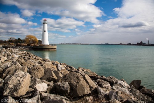A lighthouse stands on a rocky shore, with calm waters and a cloudy sky in the background.