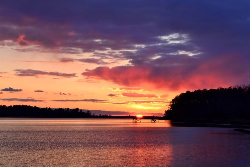 A serene lake at sunset, with vibrant orange and purple hues reflecting on the water and silhouetted trees in the background.