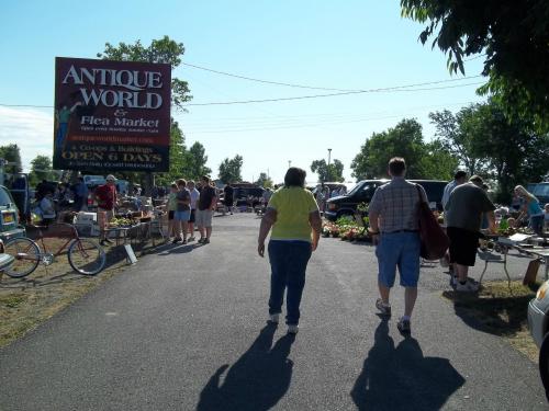 A busy flea market scene with shoppers browsing stalls under a clear blue sky, featuring a sign for Antique World.
