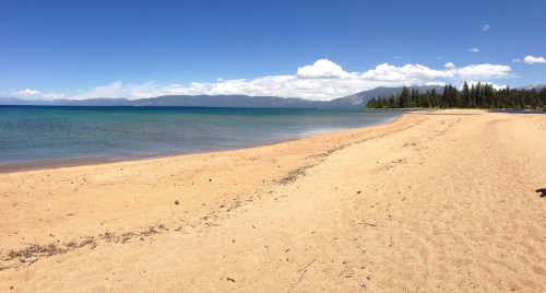 A sandy beach with clear blue water, mountains in the distance, and a bright blue sky with scattered clouds.