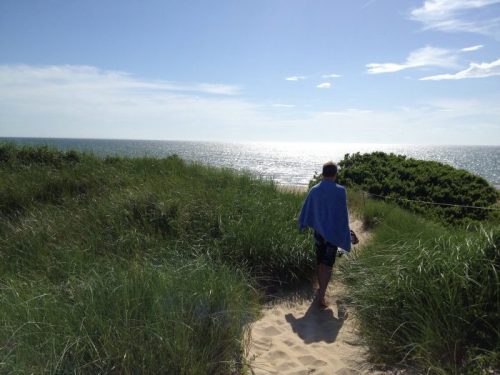 A person walks along a sandy path through tall grass, heading towards the ocean under a clear blue sky.