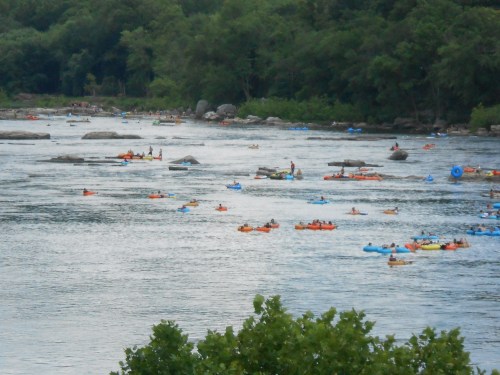 A river scene filled with people in colorful kayaks and tubes, surrounded by greenery and rocky outcrops.