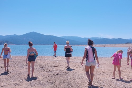 A group of people walking along a sandy shore by a lake, with mountains in the background under a clear blue sky.