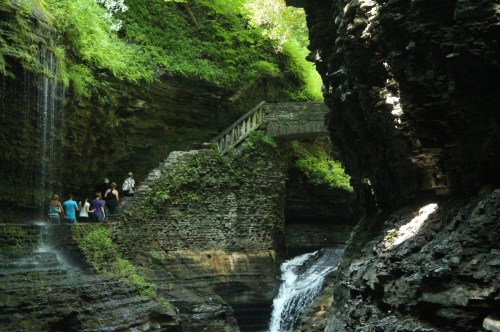 A scenic gorge with waterfalls, lush greenery, and a wooden bridge, with people exploring the natural landscape.