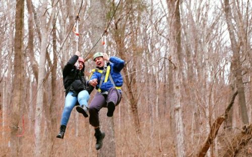Two people ziplining through a forest, smiling and enjoying the thrill of the ride. Trees surround them.