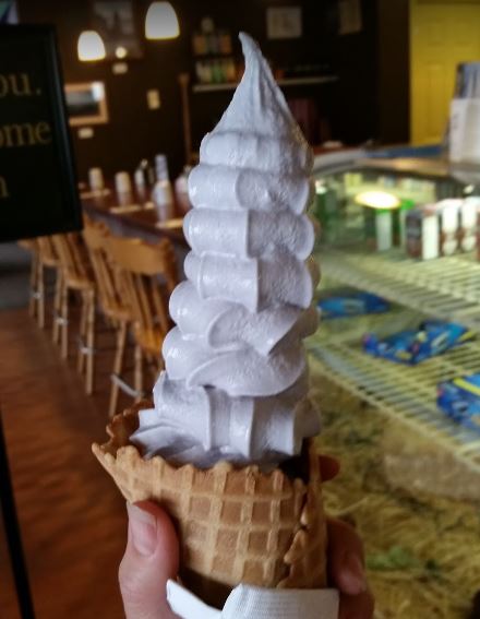 A hand holding a tall, swirled soft serve ice cream cone in a waffle cone, with a café in the background.