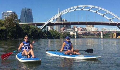 Two women paddleboarding on a river with a city skyline and bridge in the background under a clear blue sky.