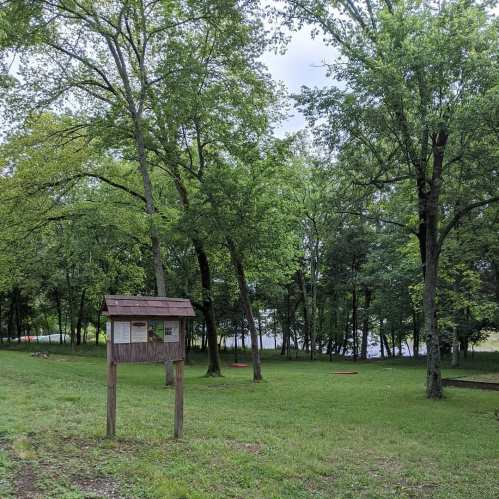 A grassy area with trees and a wooden information sign, near a calm body of water in the background.