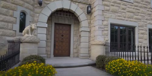 A stone house entrance with a wooden door, flanked by a lion statue and yellow flowers in the foreground.