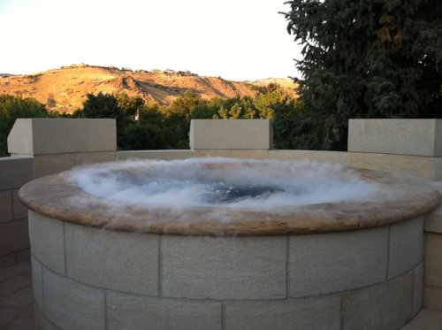 A hot tub with steam rising, set against a backdrop of hills and trees under a clear sky.