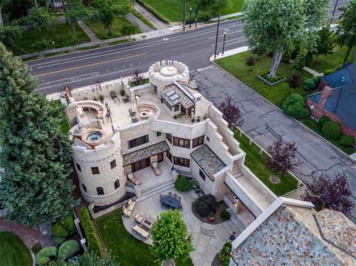 Aerial view of a unique castle-like home with a patio, surrounded by greenery and a quiet street.